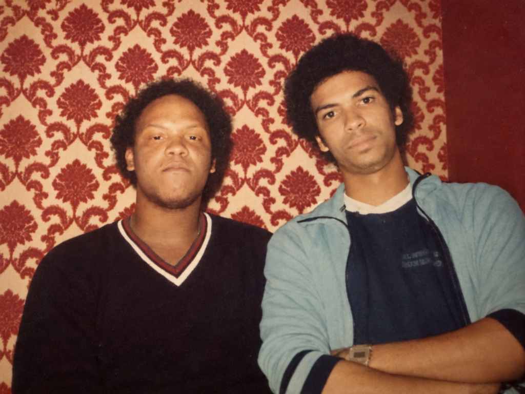 Gregory L. Hall, MD (right) and his late brother (left) as young men, posing in front of patterned red wallpaper