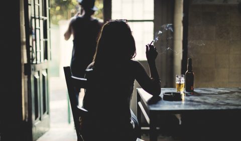 Rear view silhouette of woman sitting indoors at a table, smoking cigarette, beer glass and bottle