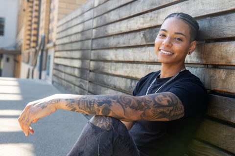 Young black woman with tattoos sits against wood wall.